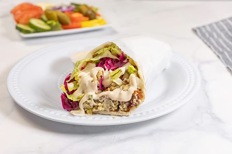 A falafel wrap with lettuce, red cabbage, and tahini sauce on a white plate with a side salad.