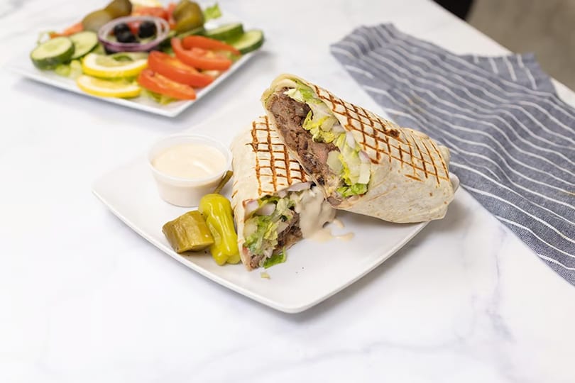 Grilled wrap with meat, lettuce, and sauce, served with pickles and a side salad.