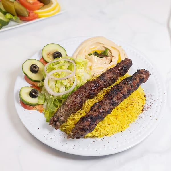 Two grilled meat skewers on yellow rice with hummus, salad, and a side of pickles and tomatoes.