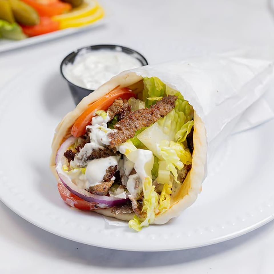 Gyro sandwich with meat, lettuce, tomatoes, onions, and white sauce, served with a side of tzatziki.