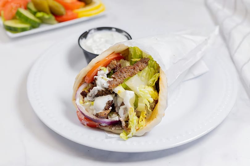 Gyro sandwich with meat, lettuce, tomatoes, onions, and white sauce, served with a side of tzatziki.