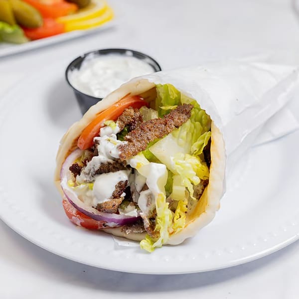 Gyro sandwich with meat, lettuce, tomatoes, onions, and white sauce, with a side of tzatziki.